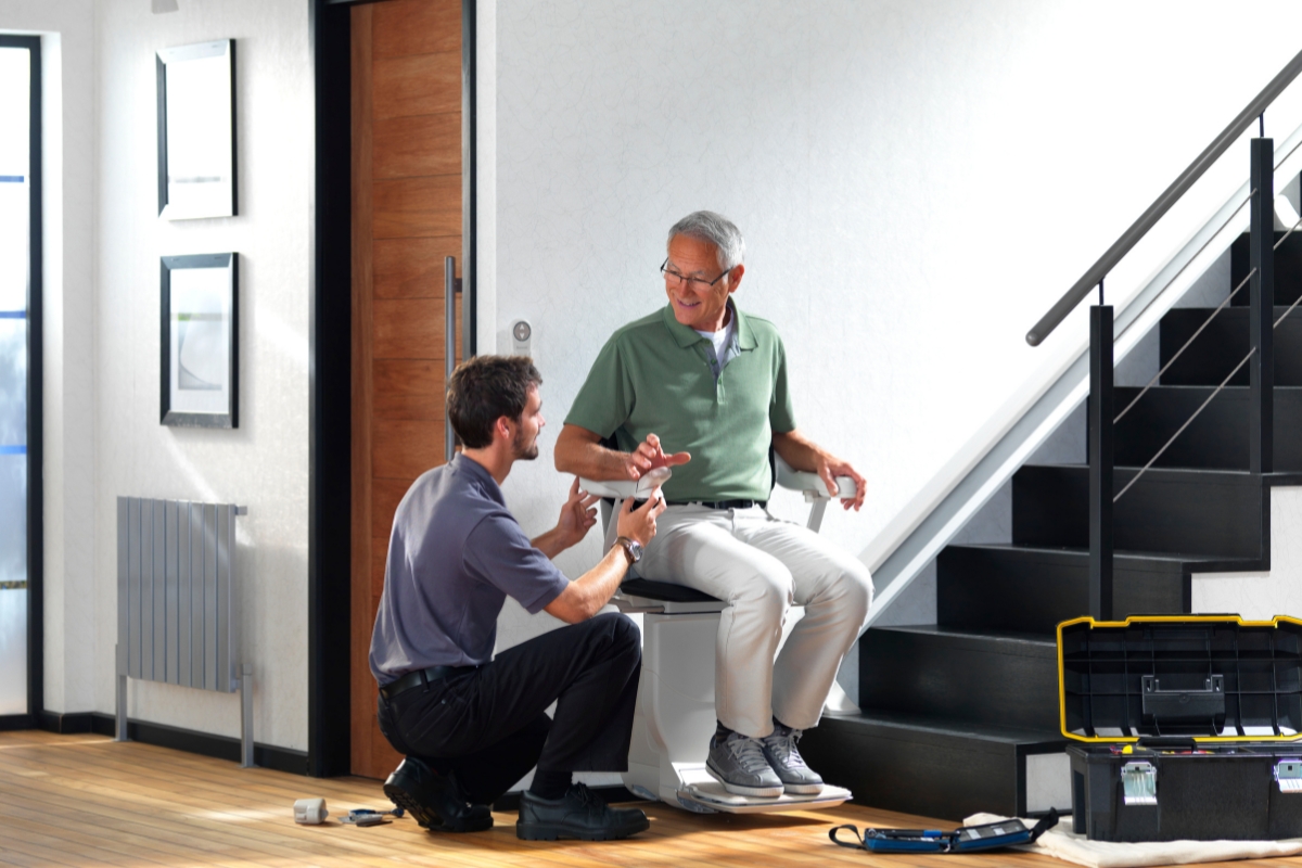 Technician kneeling beside a stairlift, explaining the controls to a smiling older man seated on the chair, with tools on the floor.