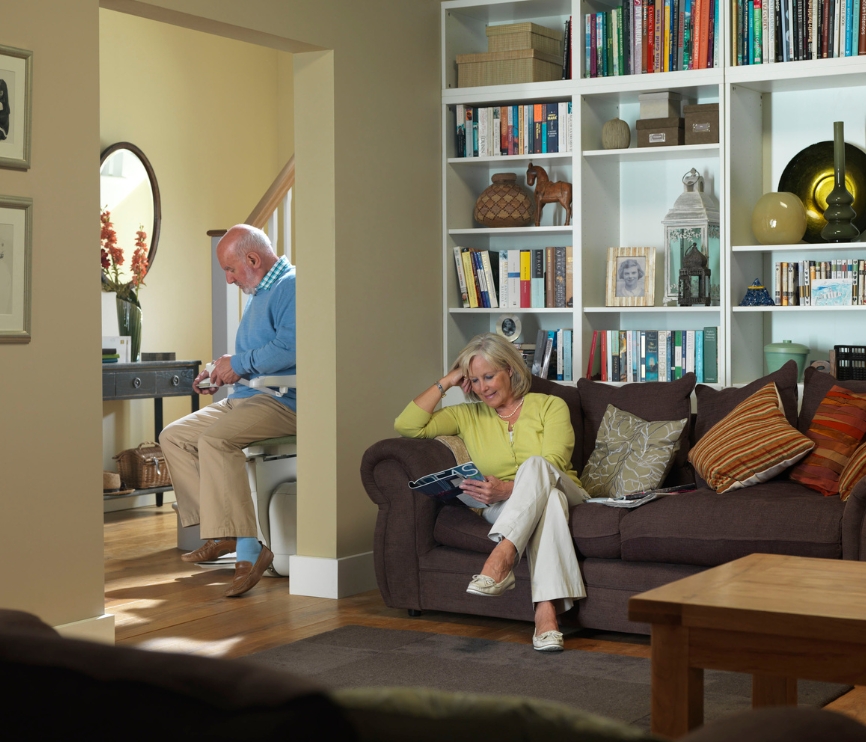 Older man using a stairlift in the hallway as an older woman reads on a sofa in a cosy living room.